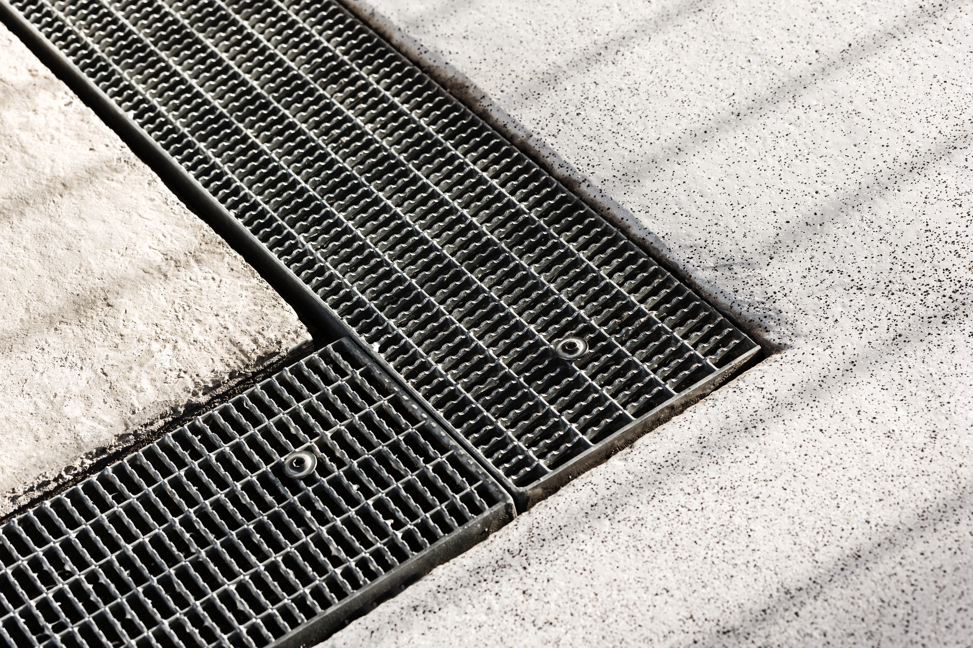 Metal drainage grates set into a concrete surface, with visible bolts securing the grates in place. The concrete shows some texture and speckles.