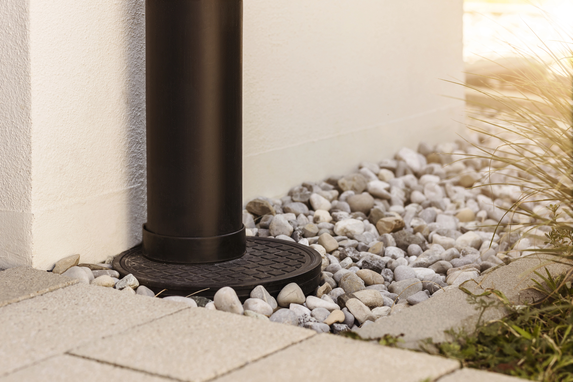 Black drainage pipe terminates at a round drain cover, surrounded by white decorative stones and tan paving slabs beside a white building exterior.