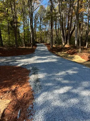 A newly constructed gravel road curves through a wooded area with trees on both sides and patches of red soil visible along the edges.