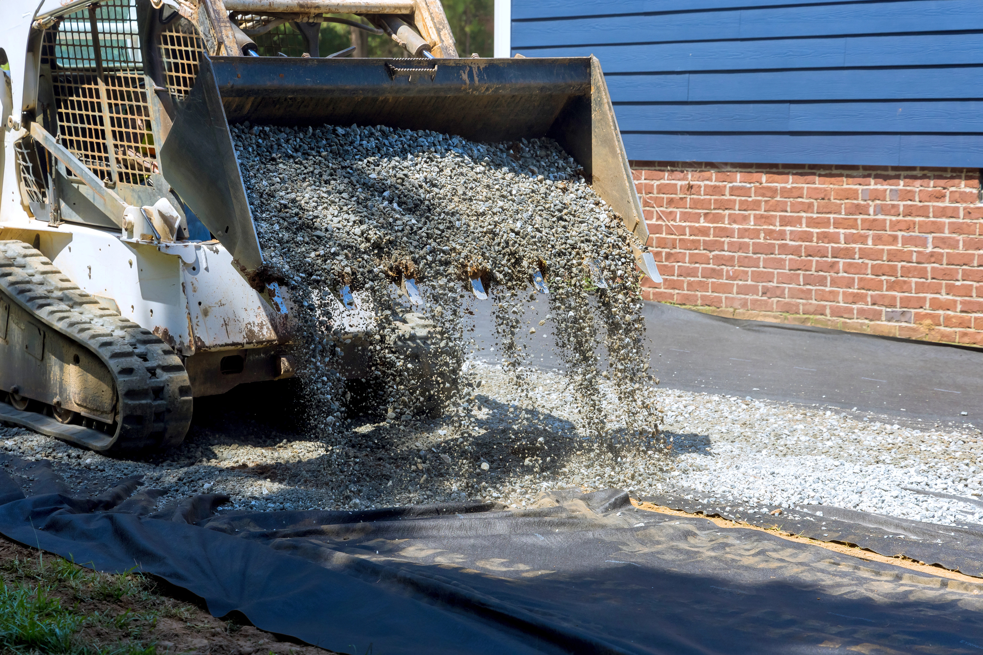 A skid steer loader pours gravel onto a black geotextile fabric on a driveway beside a brick and blue-sided house.