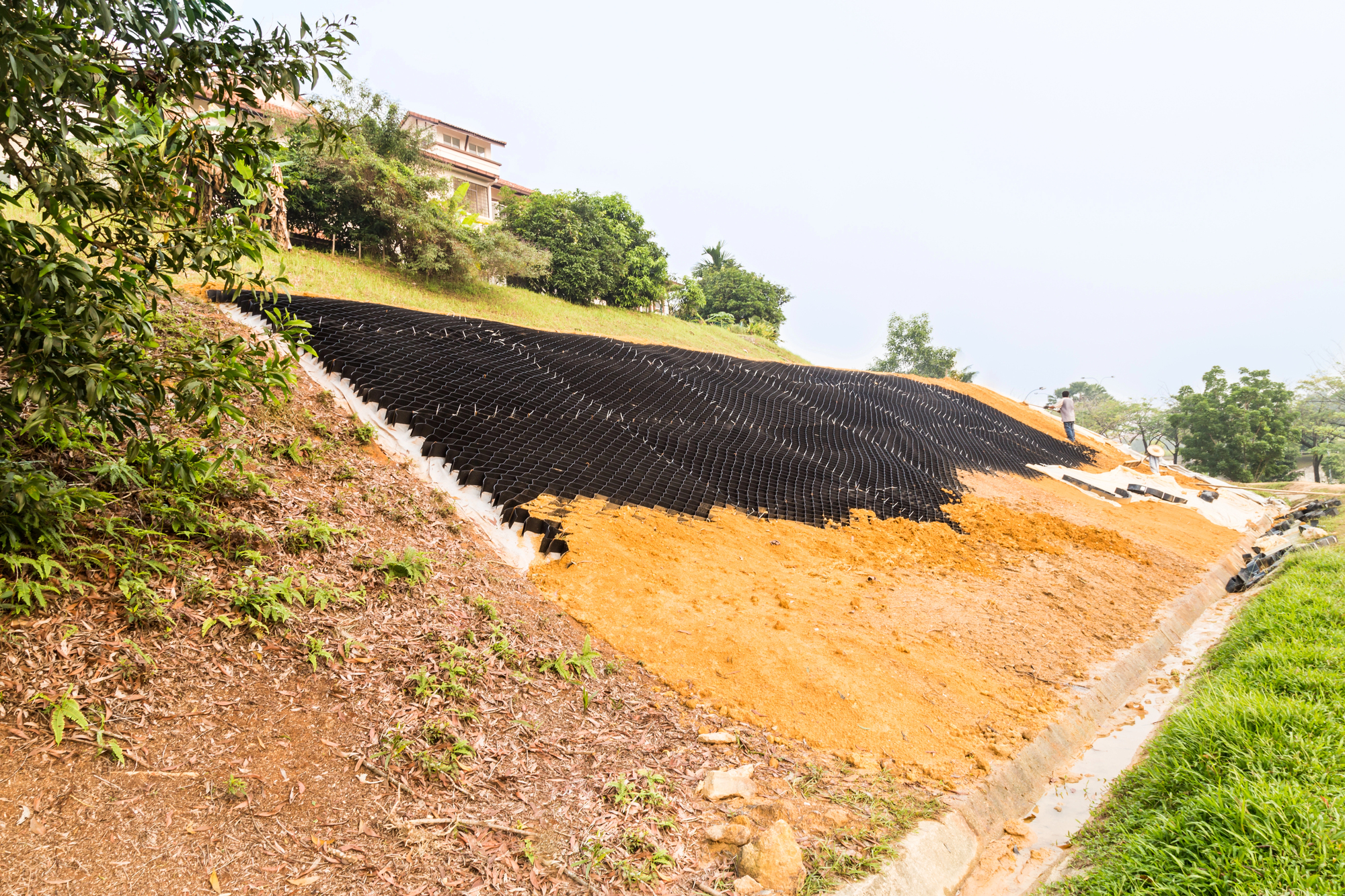 A hillside is being stabilized with black geocell mats and soil. The slope appears partially covered, with vegetation and a house visible at the top.