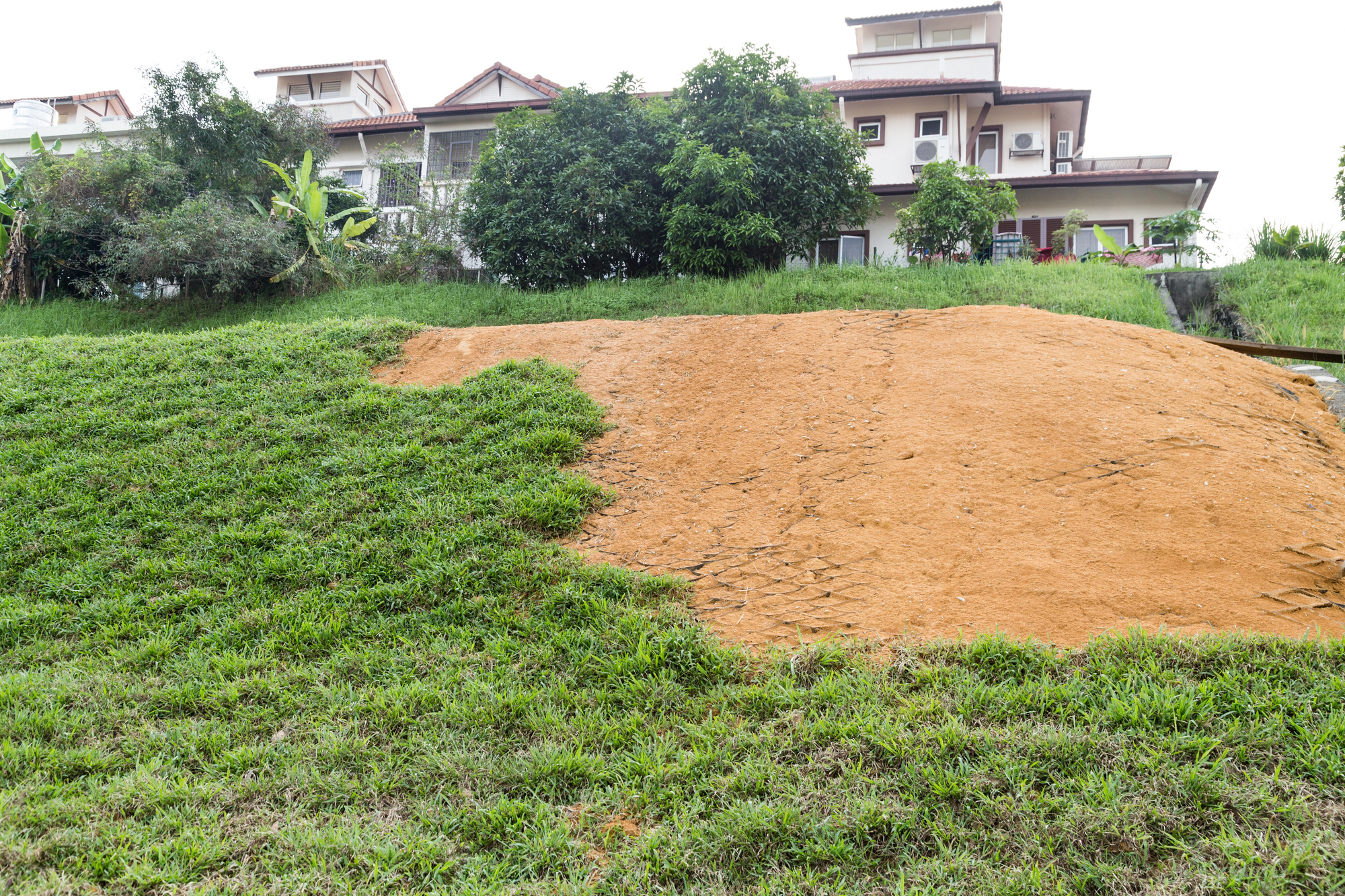 A patch of bare soil with tire tracks is surrounded by grassy areas, in front of residential houses with trees in the background.