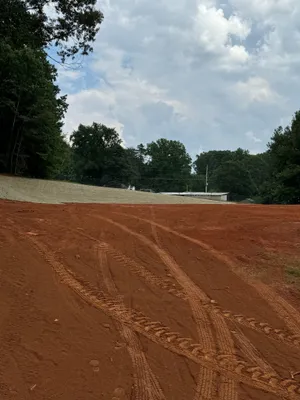 Tire tracks on reddish dirt lead toward a small building and trees under a partly cloudy sky.