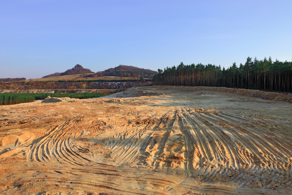 A wide sandy area with tire tracks, bordered by a forest on the right and hills in the background under a clear sky.