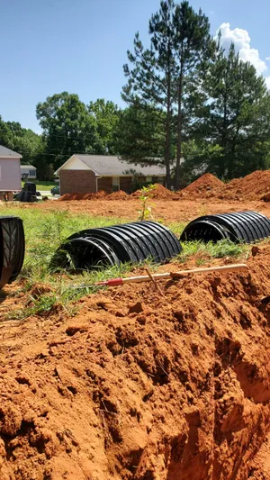 Large black plastic septic drain pipes partially buried in a red dirt construction site, with trees and houses visible in the background.