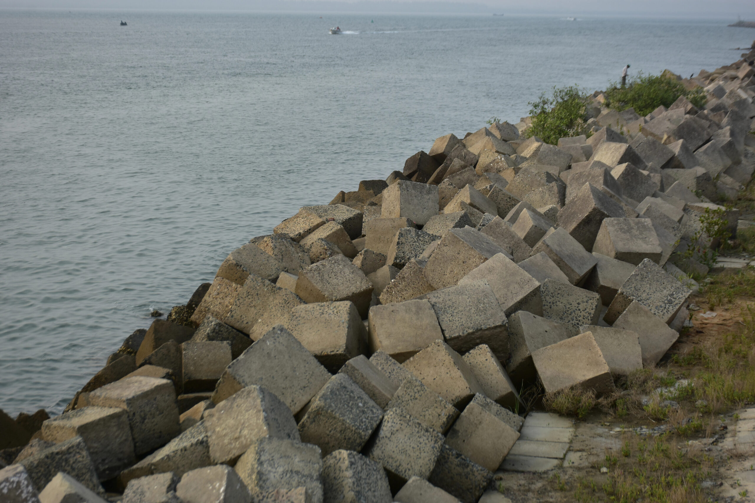 Large concrete blocks form a sloped barrier along the shoreline, with calm sea water in the background and some greenery on the right side.