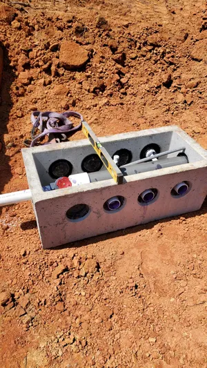 A concrete utility box with multiple conduit openings sits on red soil, containing cables, a level tool, and a strap placed on top.